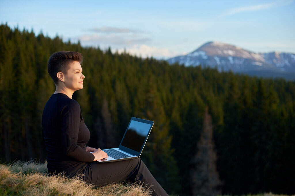 happy young woman working on her laptop outdoors enjoying view of mountains